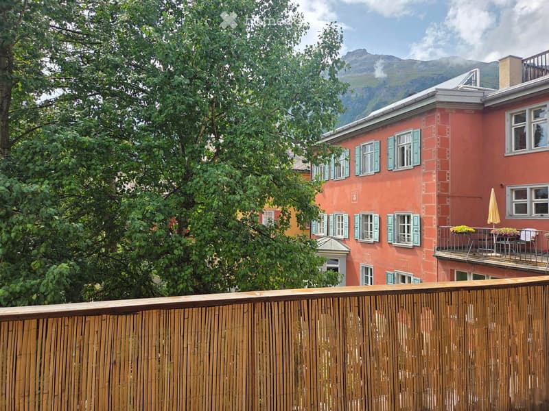 A multi-story red building with white-framed windows and green shutters, featuring a balcony with outdoor furniture and a scenic mountain view in the background.