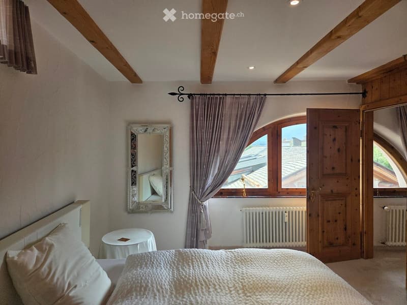 Cozy attic bedroom with wooden beams, large window with view, bed, mirror, small round table, and radiator.