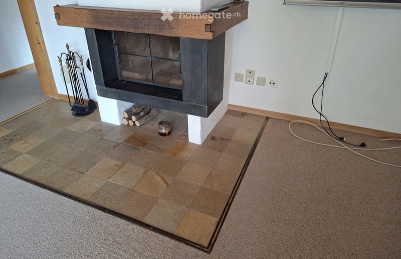 A modern fireplace with a wooden mantelpiece, surrounded by tiled and carpeted flooring, with fireplace tools and logs nearby.