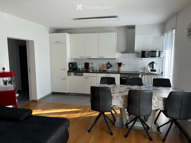 Modern kitchen and living area with white cabinets, a dining table with chairs, a sofa, and various kitchen appliances.