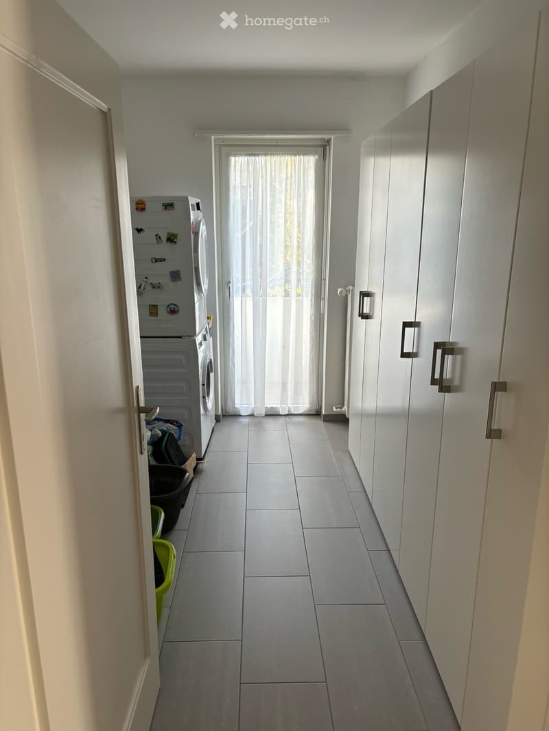 A narrow hallway with tiled flooring, white walls, and built-in storage units. A washing machine and dryer are positioned on the left side, with a window at the end covered by sheer curtains.