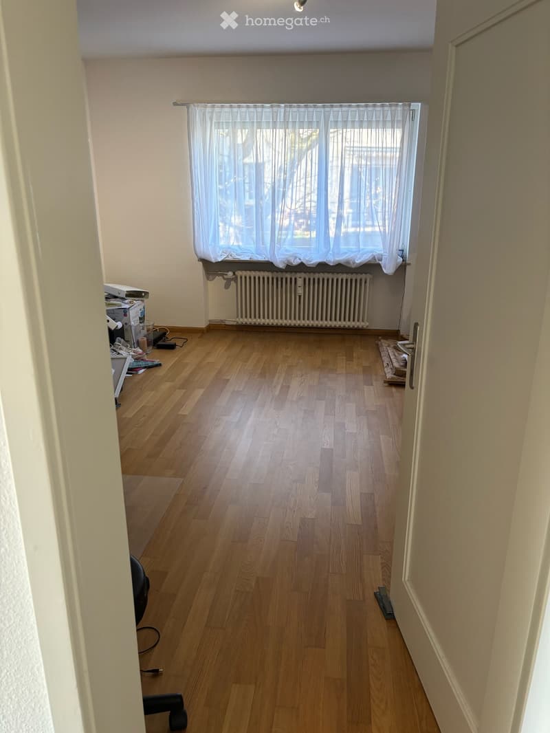 Empty room with wooden flooring, white walls, a window with white curtains, and a radiator below the window. No furniture or equipment is present.