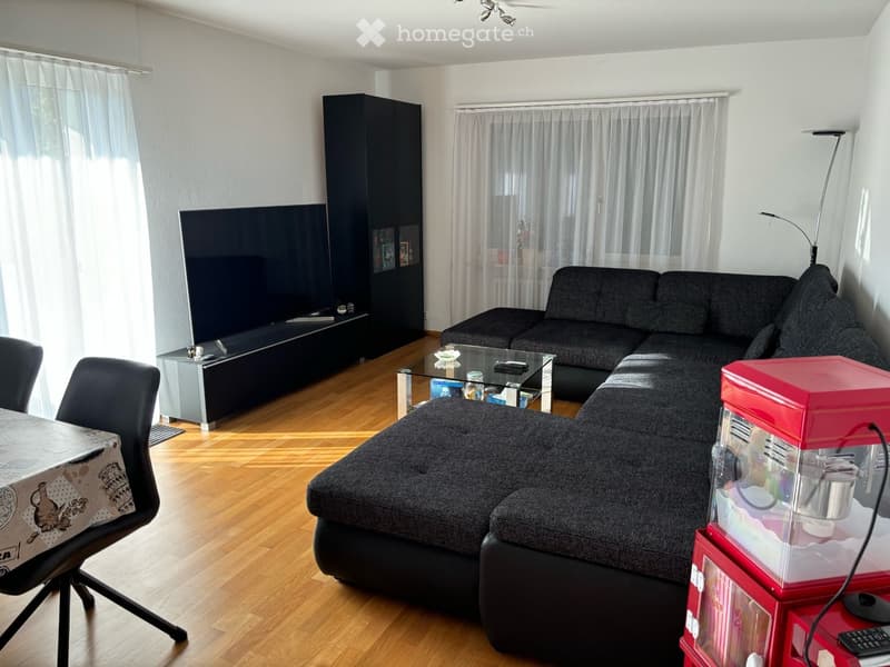 Modern living room with a grey sectional sofa, glass coffee table, wooden floor, TV, and a small table with chairs.