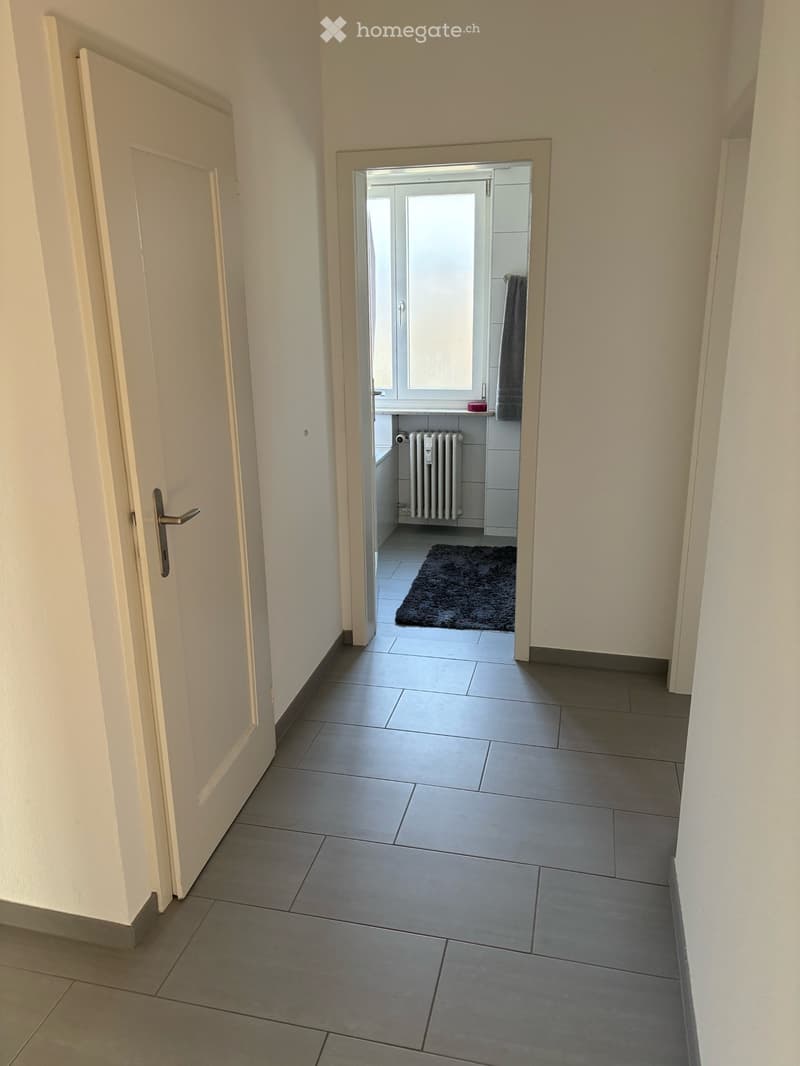 A clean, tiled hallway with a door on the left leading to a bathroom. The bathroom has a window, a radiator, a towel, and a dark rug on the floor.