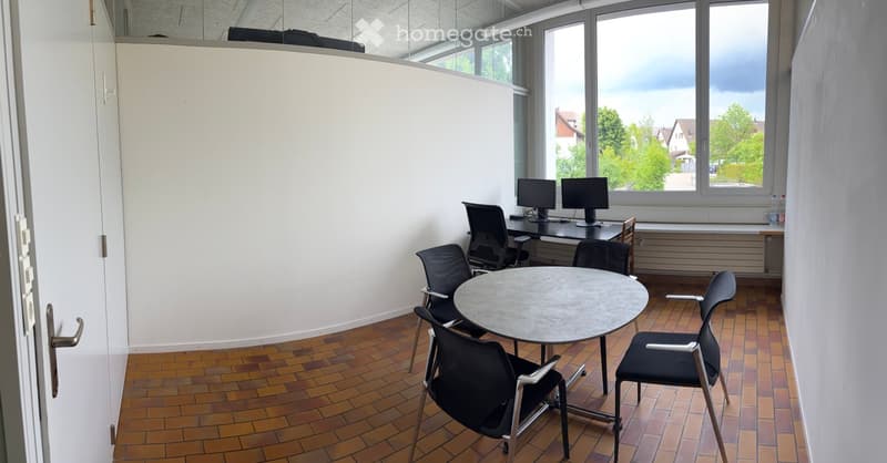 Office space with a round table, four chairs, two computer setups with monitors, and large windows overlooking a green area.