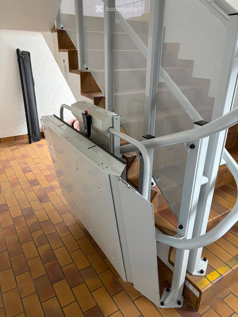 A modern staircase with a metal railing and a grey lift platform, featuring a tiled floor and white walls.