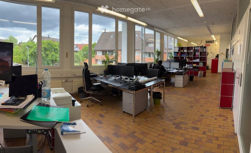 Office space with desks, chairs, computers, and office supplies. Large windows provide natural light, and the floor is tiled.