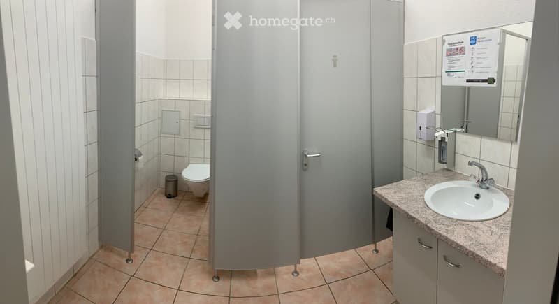Public restroom with a toilet, sink, mirror, hand sanitizer dispenser, paper towel dispenser, and a trash can. The walls are tiled, and the floor is tiled with a light brown color.