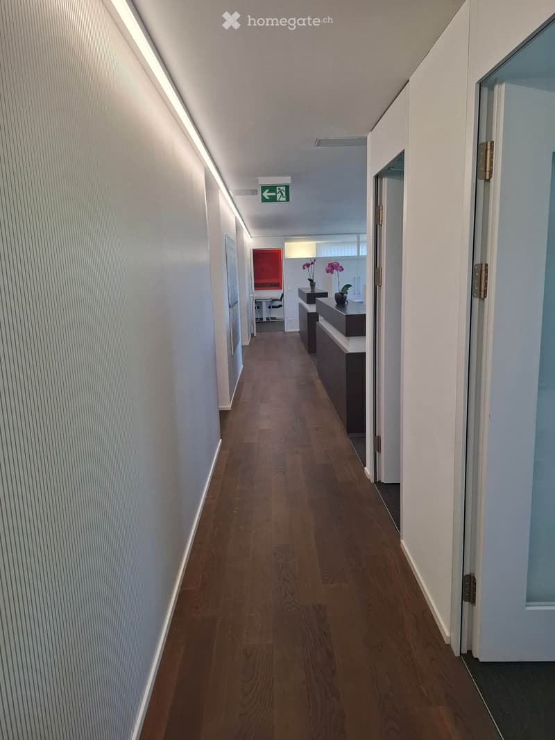 Modern hallway with wooden flooring, white walls, and recessed lighting. Features an emergency exit sign and a small reception area visible at the end.