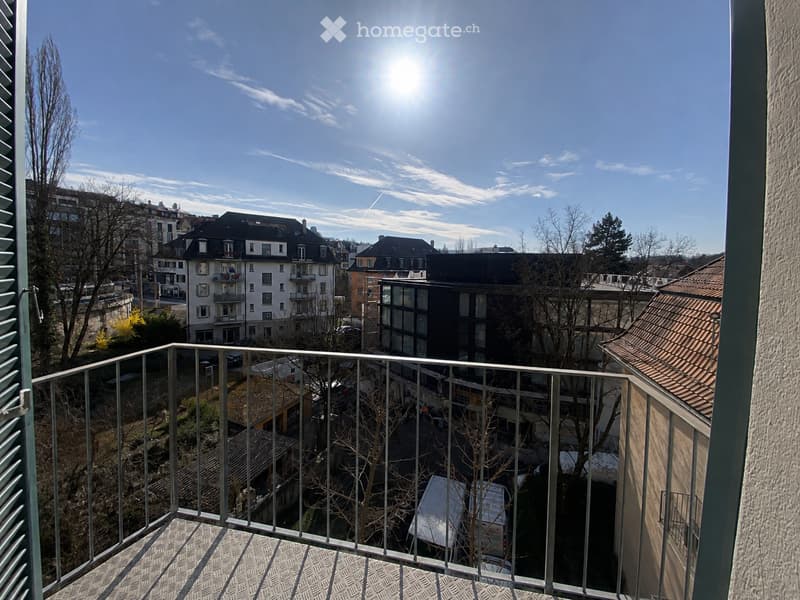Balcony with metal railing overlooking a cityscape of buildings, parked cars, and trees under a clear blue sky with the sun shining brightly.