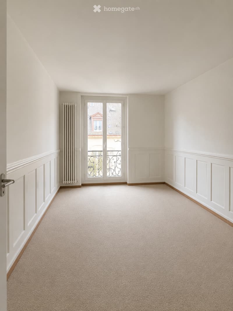 Empty room with beige carpet, white walls, a door, and a large window with a view of a building outside.