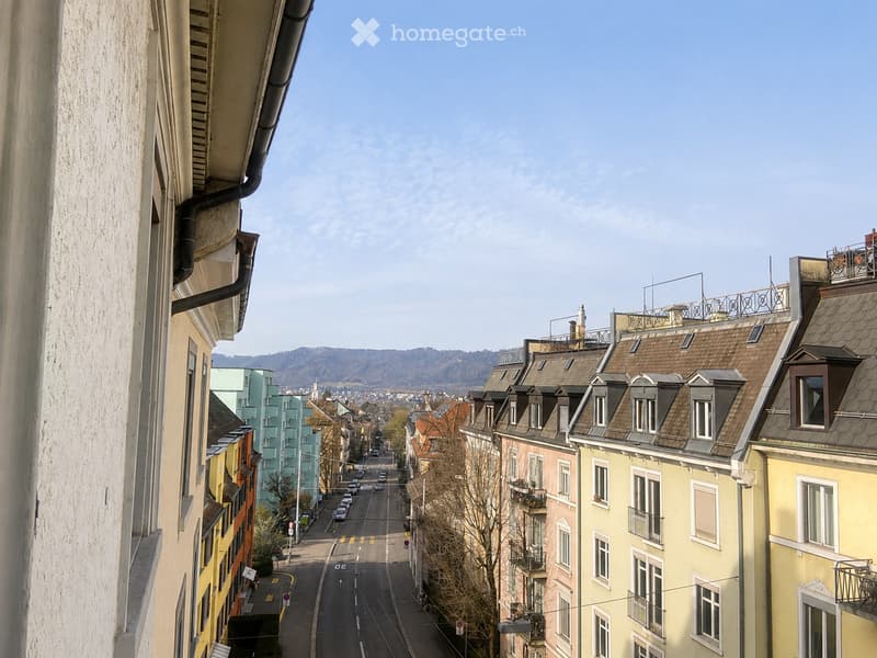 A view of a city street lined with colorful residential buildings, featuring balconies and varied architectural styles, with a clear sky and distant hills in the background.