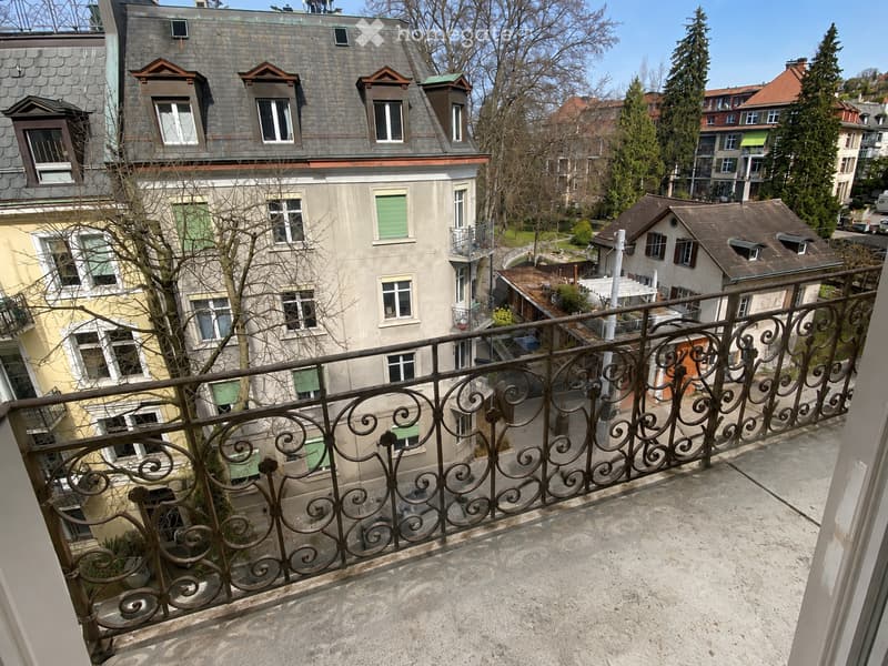 Balcony with ornate metal railing overlooking a residential area with multiple buildings, trees, and a clear blue sky.