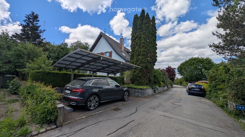 A two-story house with a white exterior and a gray roof, featuring a covered parking area for a car. The driveway is bordered by greenery and trees, with a clear blue sky overhead.