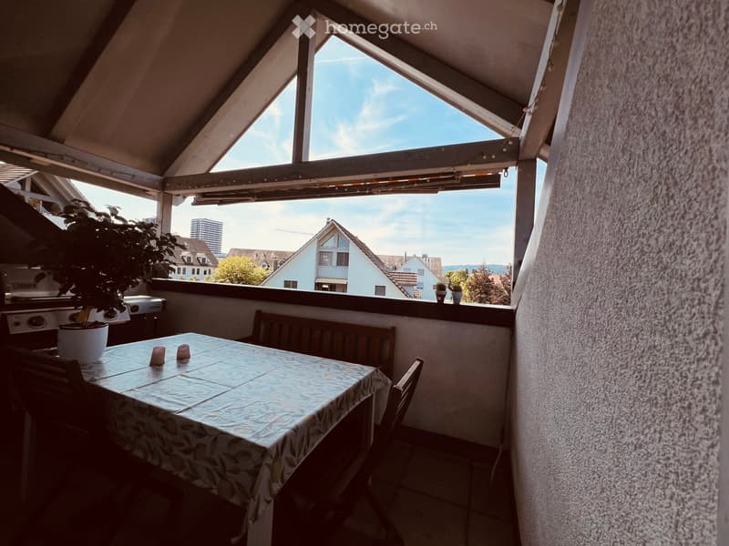 Balcony with a table, chairs, bench, and potted plant, overlooking a cityscape with houses and a clear sky.