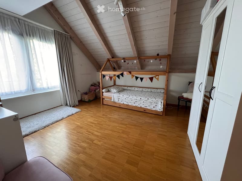 Cozy attic bedroom with wooden slanted ceilings, a lofted twin-sized bed, a small stool, a mirror, and a wardrobe. The room features wooden flooring, a white shag rug, and a window with curtains.