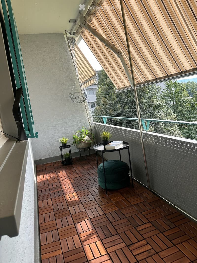 Balcony with wooden decking, glass railing, and striped awning. Includes a green stool, small table with potted plants and a book.