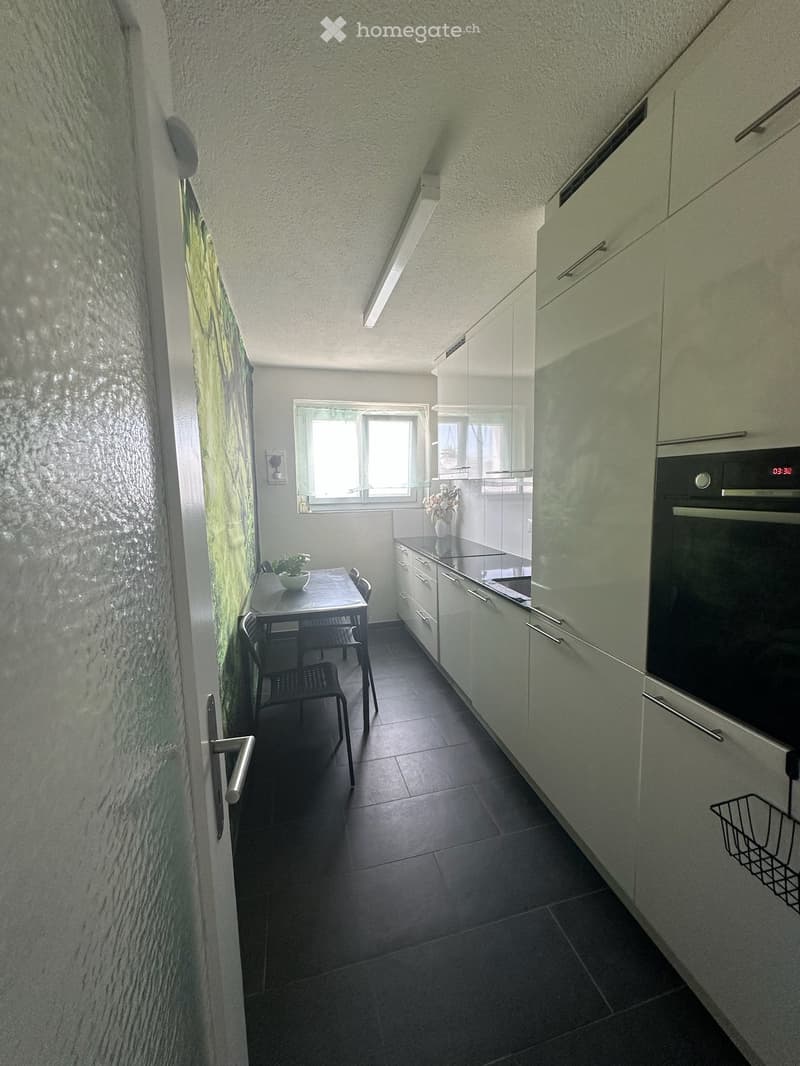 Modern kitchen with white cabinets, dark tiled floor, and a small dining area with a table and chairs. Features an oven, a vase with flowers, and a potted plant on the table.