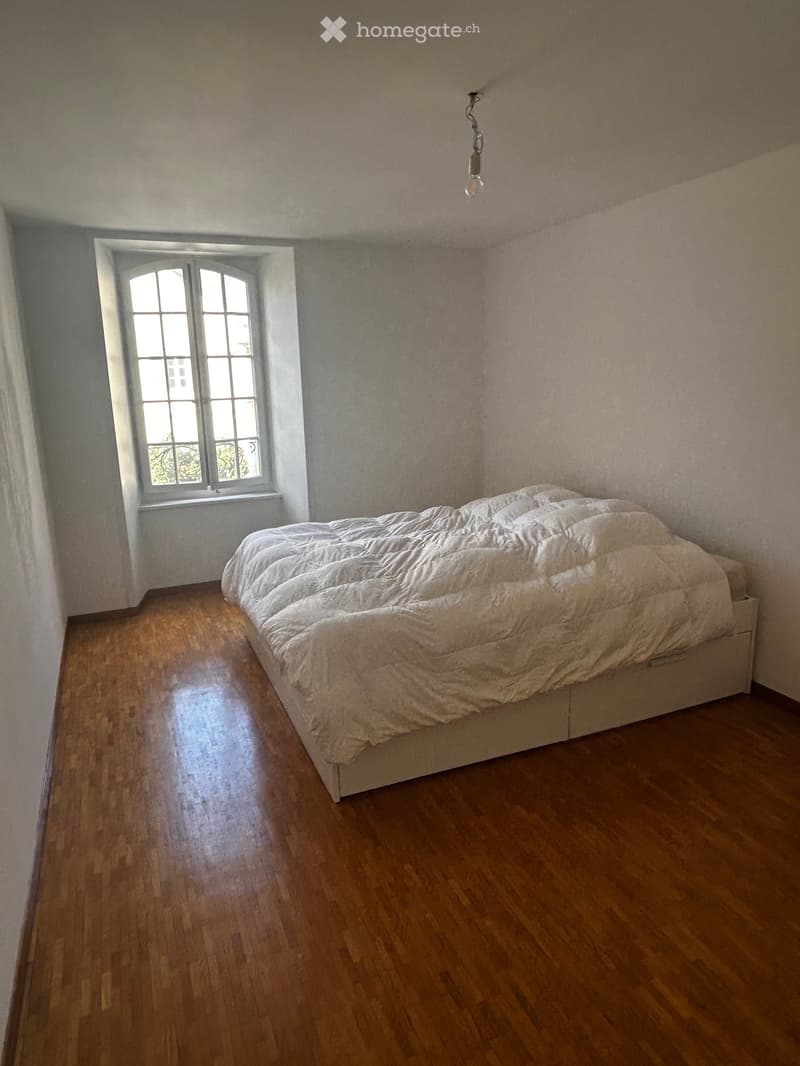 Bedroom with wooden floor, large window, and a neatly made bed with white bedding.