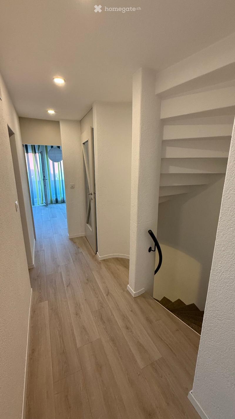 A narrow hallway with wooden flooring, white walls, and recessed ceiling lights. A staircase is visible on the right, leading upwards, with a handrail and open shelving. An open doorway on the left leads to another room with large windows and a visible ha