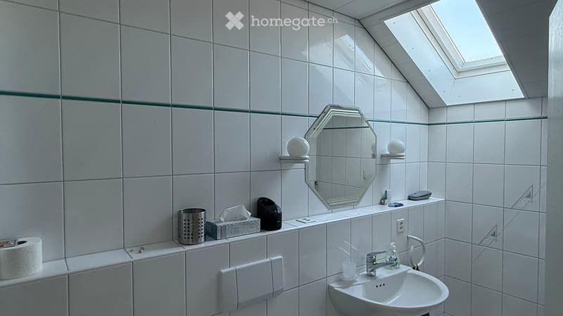 Modern bathroom with white tiled walls and a skylight window. Features a sink with a modern faucet, a large octagonal mirror with shelves, and various toiletries neatly arranged on the counter.