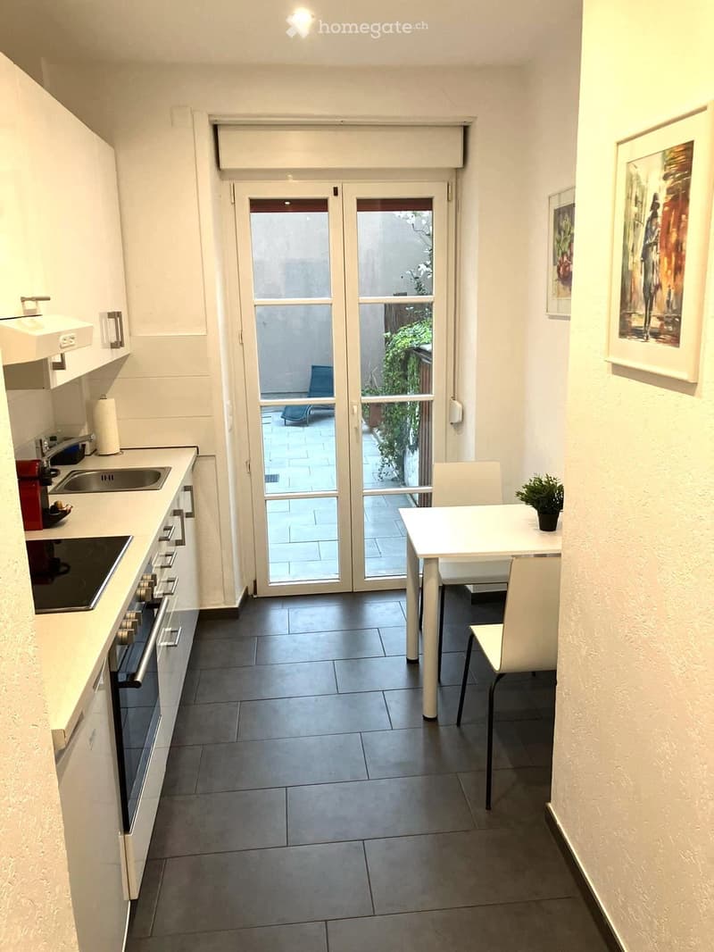 Modern kitchen with white cabinets, stainless steel appliances, and a small dining area with a table and chairs. The space features a large glass door leading to a balcony with outdoor seating.