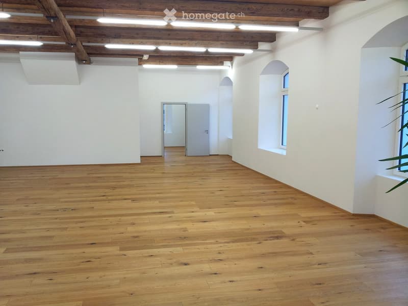 Empty room with wooden flooring, white walls, exposed wooden ceiling beams, and arched windows.