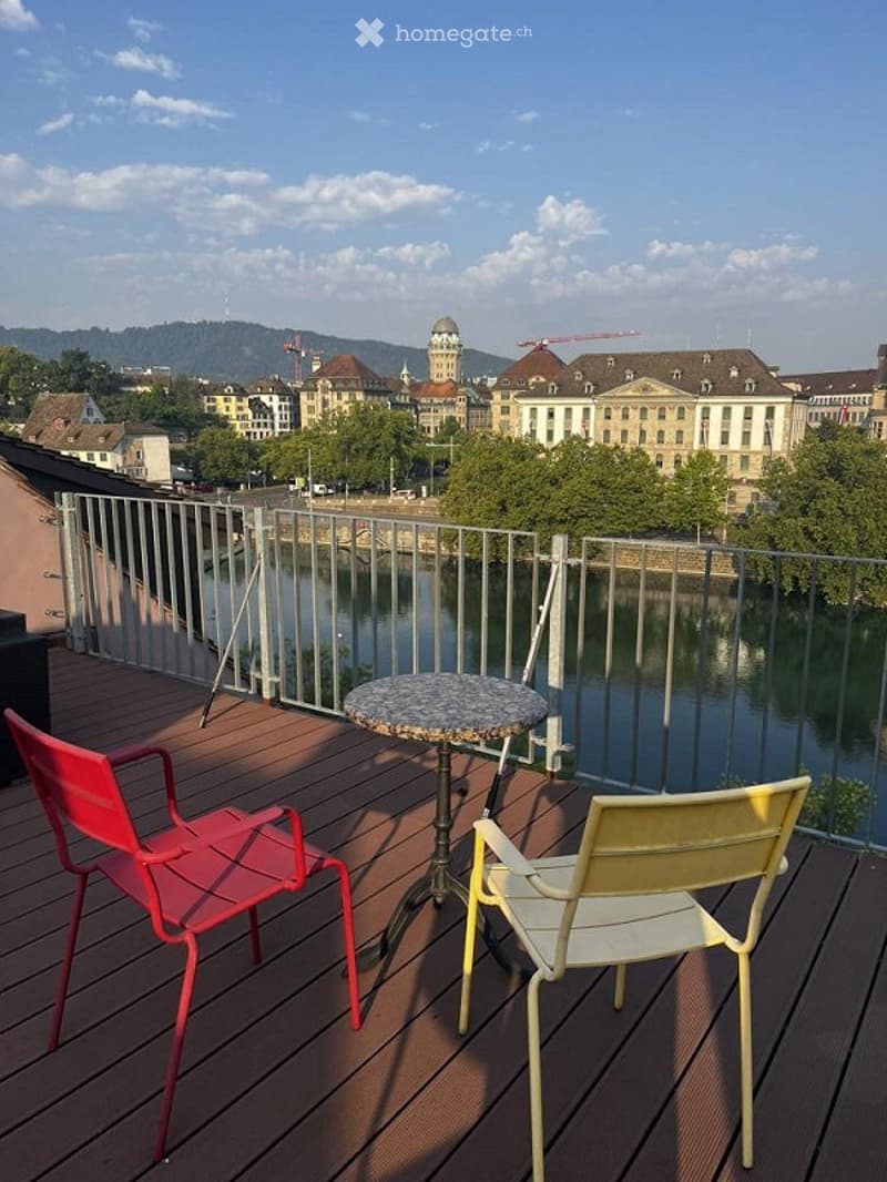 Balcony with wooden decking, metal railing, round table, and two chairs overlooking a river and cityscape.