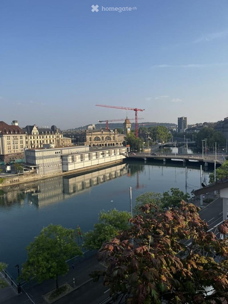 A scenic view from a balcony or terrace overlooking a river with reflections of buildings, a bridge, and construction cranes in the background.