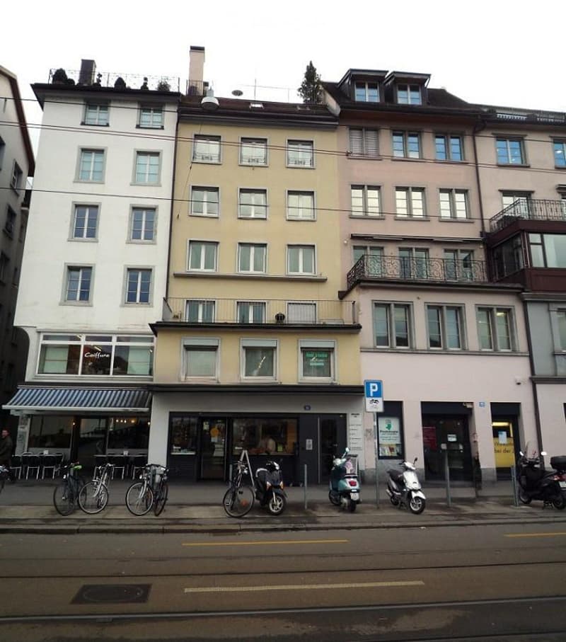 Multi-story urban building with a mix of residential and commercial spaces, featuring balconies, bicycles, and scooters parked on the street.