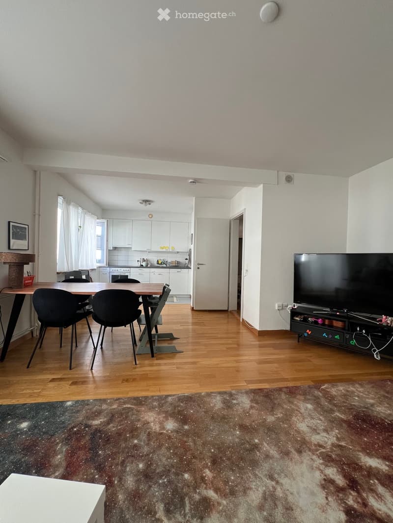 Open-plan living and dining area with wooden flooring, a large flat-screen TV on a stand, a dining table with chairs, and a kitchen area with white cabinets and appliances.
