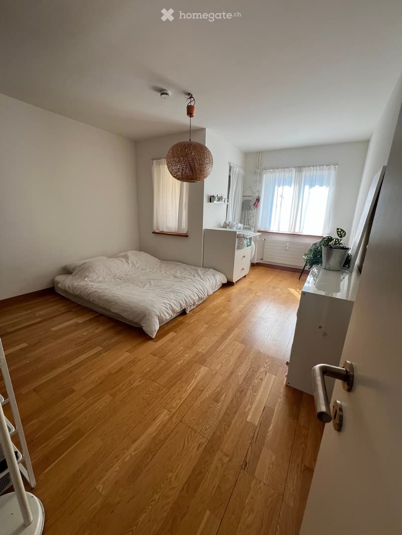 Cozy bedroom with wooden flooring, a single bed with white bedding, a nightstand with a mirror, a hanging woven lamp, and a small potted plant near the window.