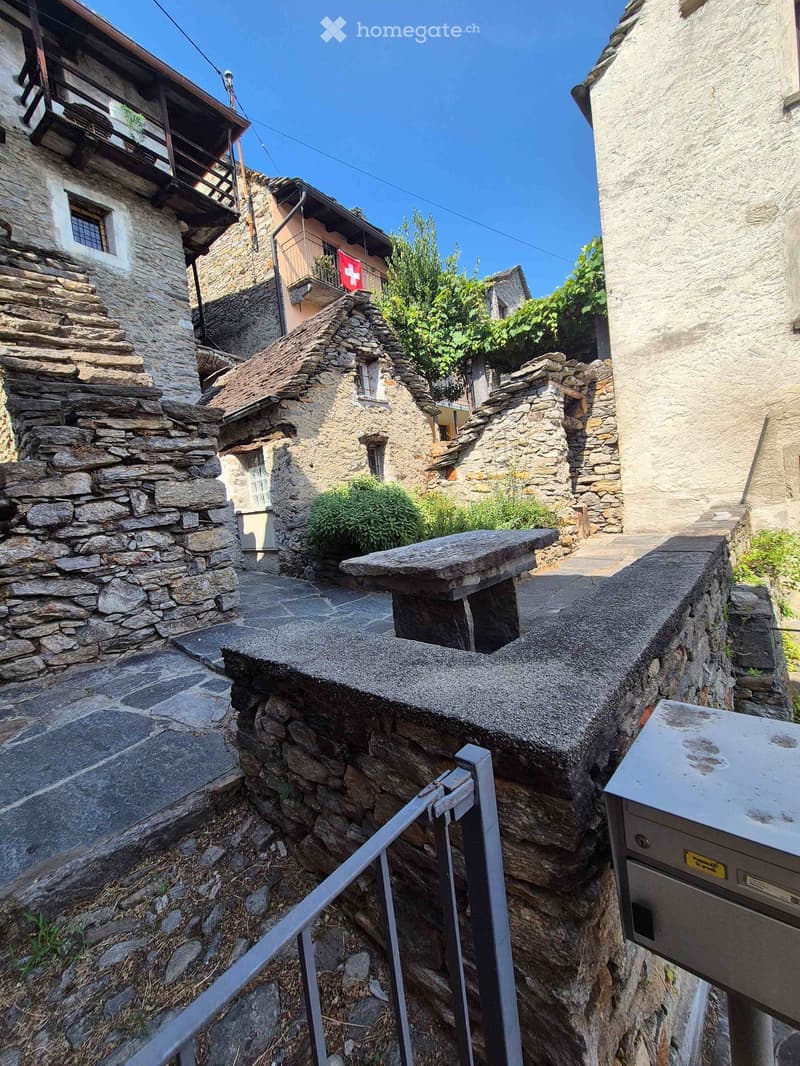 A narrow stone staircase leads up between old stone buildings with wooden balconies and small gardens, featuring a metal railing and a mailbox on the right side.