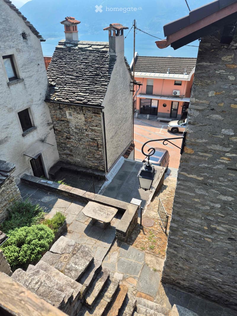 Narrow stone staircase leading up to a stone balcony overlooking a scenic view of a lake and mountains. The area includes stone walls, a small bench, and a lantern.