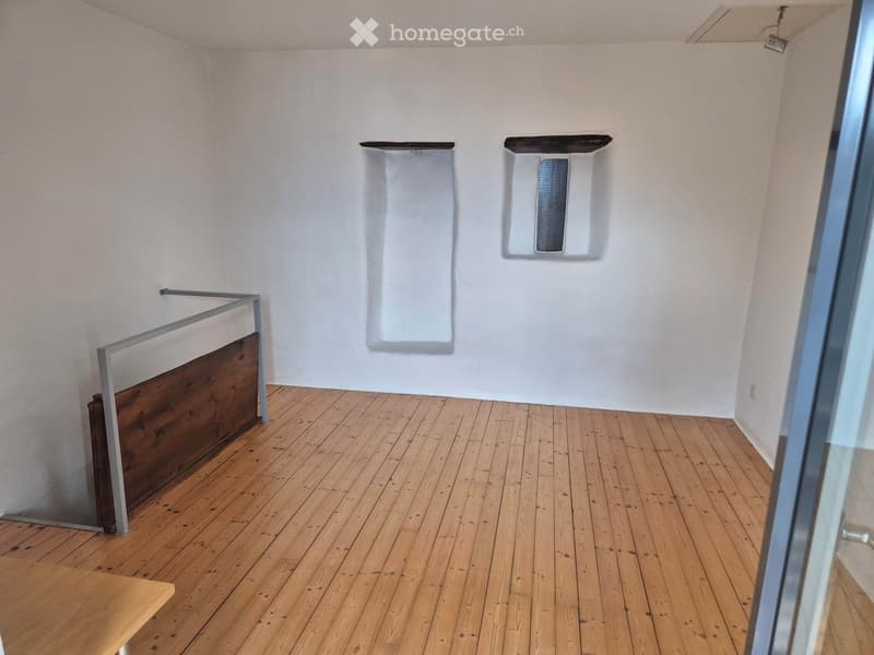 Empty room with wooden flooring, white walls, and a glass door on the right side. There are two wall-mounted white panels and a small wooden bench in the foreground.