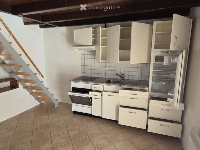 Compact kitchen with white cabinets, stainless steel appliances, and a tiled backsplash. A wooden staircase leads upwards.