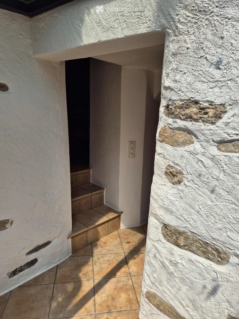 A narrow hallway with white plaster walls, a tiled floor, and a small step leading to another area. The walls show some wear and patches of exposed stone.