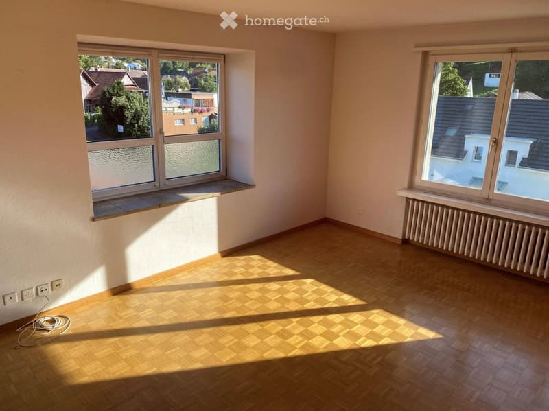 Empty room with wooden flooring, large windows, radiator, and electrical outlets. Sunlight streams in from the windows, casting shadows on the floor.