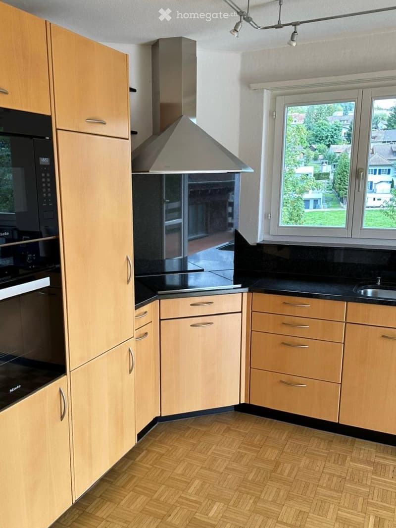 Modern kitchen with light wooden cabinets, black countertops, and stainless steel appliances including a microwave, oven, and range hood. The room features a herringbone wooden floor and large windows offering a view of the outdoors.