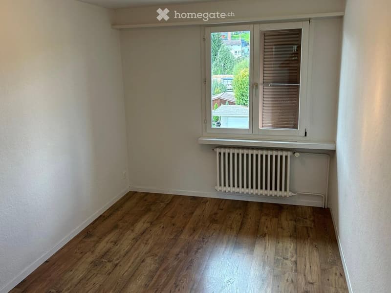 Empty room with wooden flooring, white walls, and a radiator under a window with blinds. The window offers a view of trees and neighboring houses.