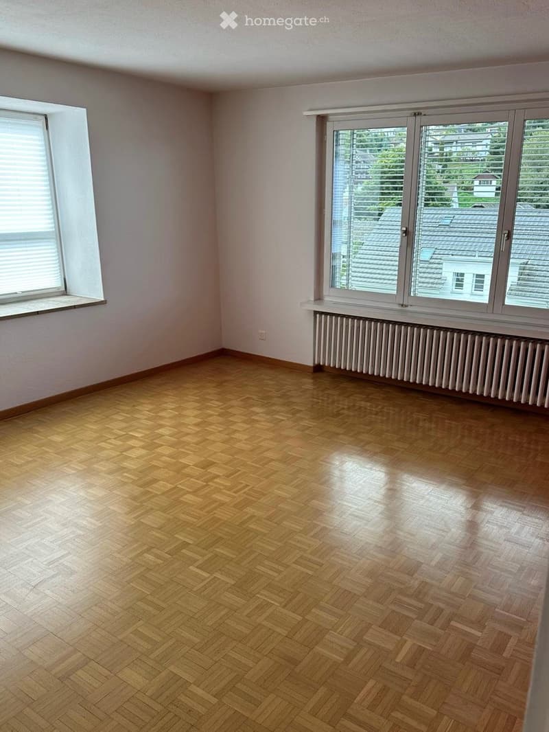 Empty room with wooden herringbone flooring, white walls, and large windows with blinds. Radiator visible under one window.