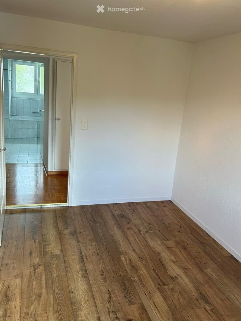 Empty room with wooden flooring and white walls, featuring a doorway leading to a tiled bathroom.