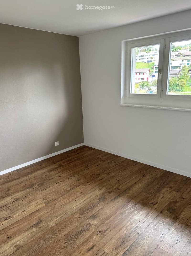 Empty room with wooden flooring, white walls, and a large window showing a view of the outside area.