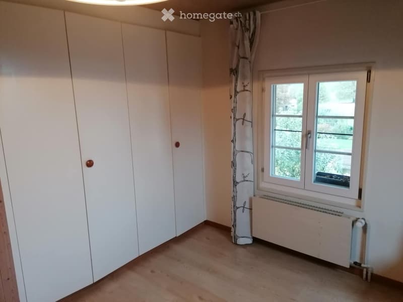 Empty room with light-colored walls, wooden flooring, large window with white frame, and white built-in wardrobe.