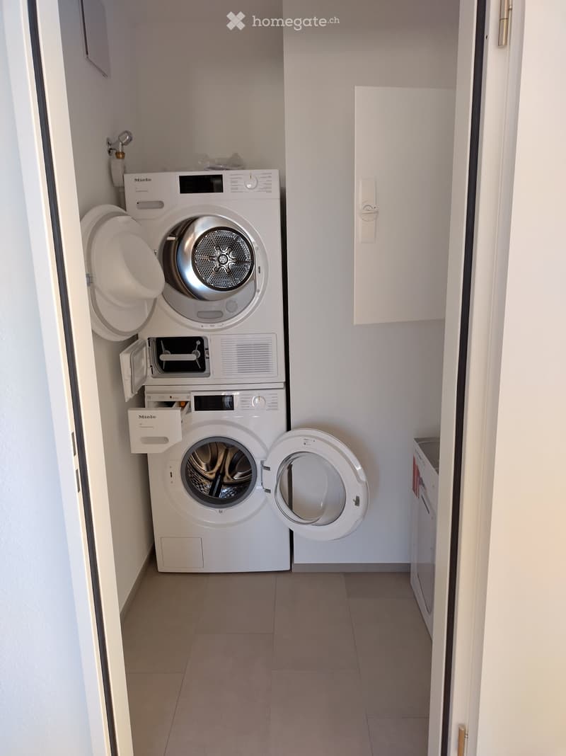 room with 2 stacked white washing machines, white cabinetry, tiled floor, small trash bin