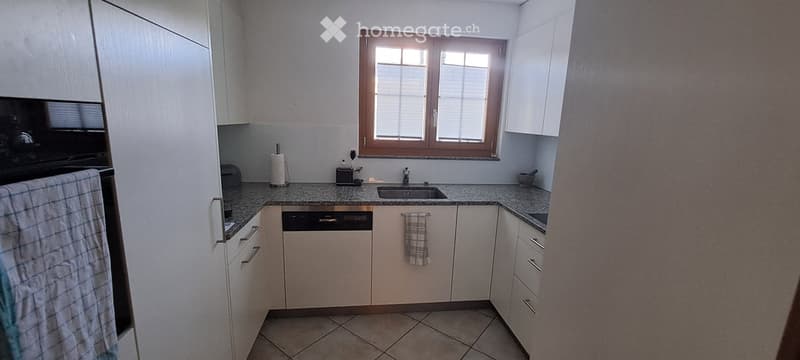 Modern kitchen with white cabinets, granite countertops, a sink, dishwasher, and oven. Features a window with blinds and tiled flooring.