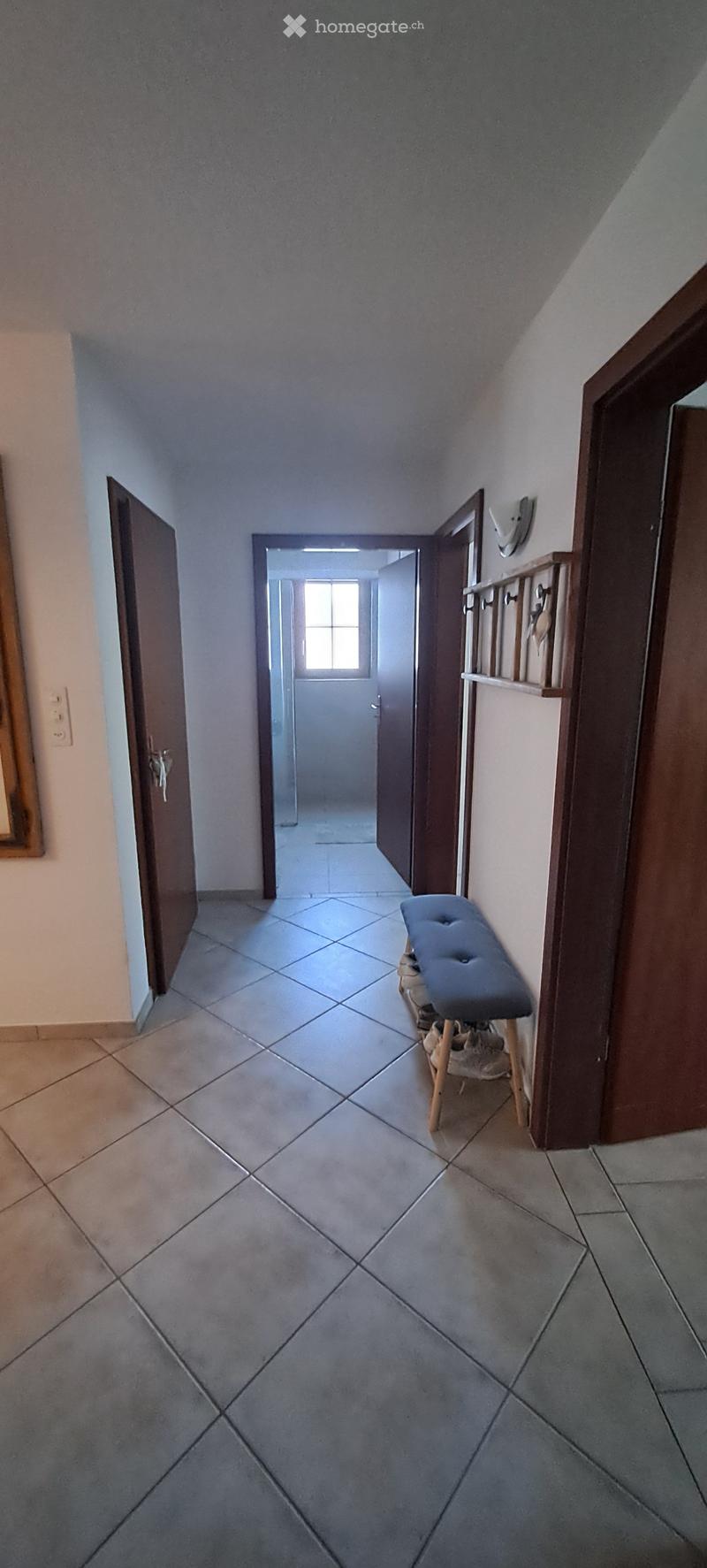 A narrow hallway with tiled flooring, several open doors leading to other rooms, and a small bench with shoes placed on it.