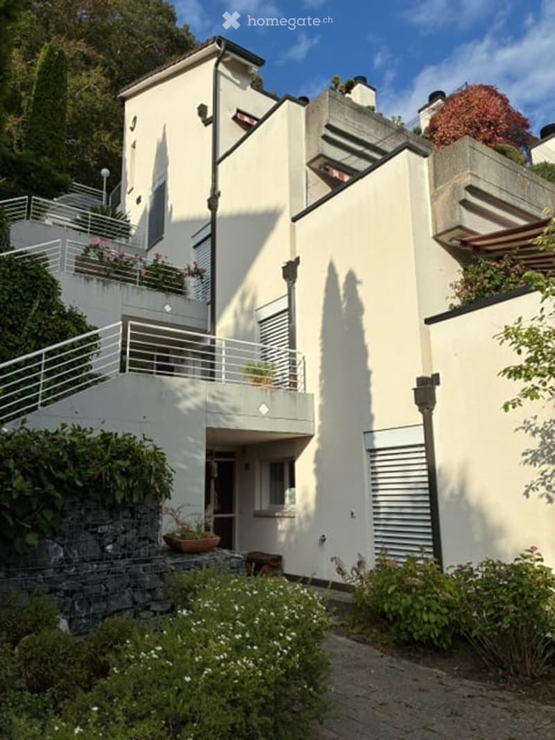 Modern two-story house with white exterior, large windows, and a balcony with metal railing. The house features a stone staircase leading to the entrance, surrounded by greenery and potted plants.