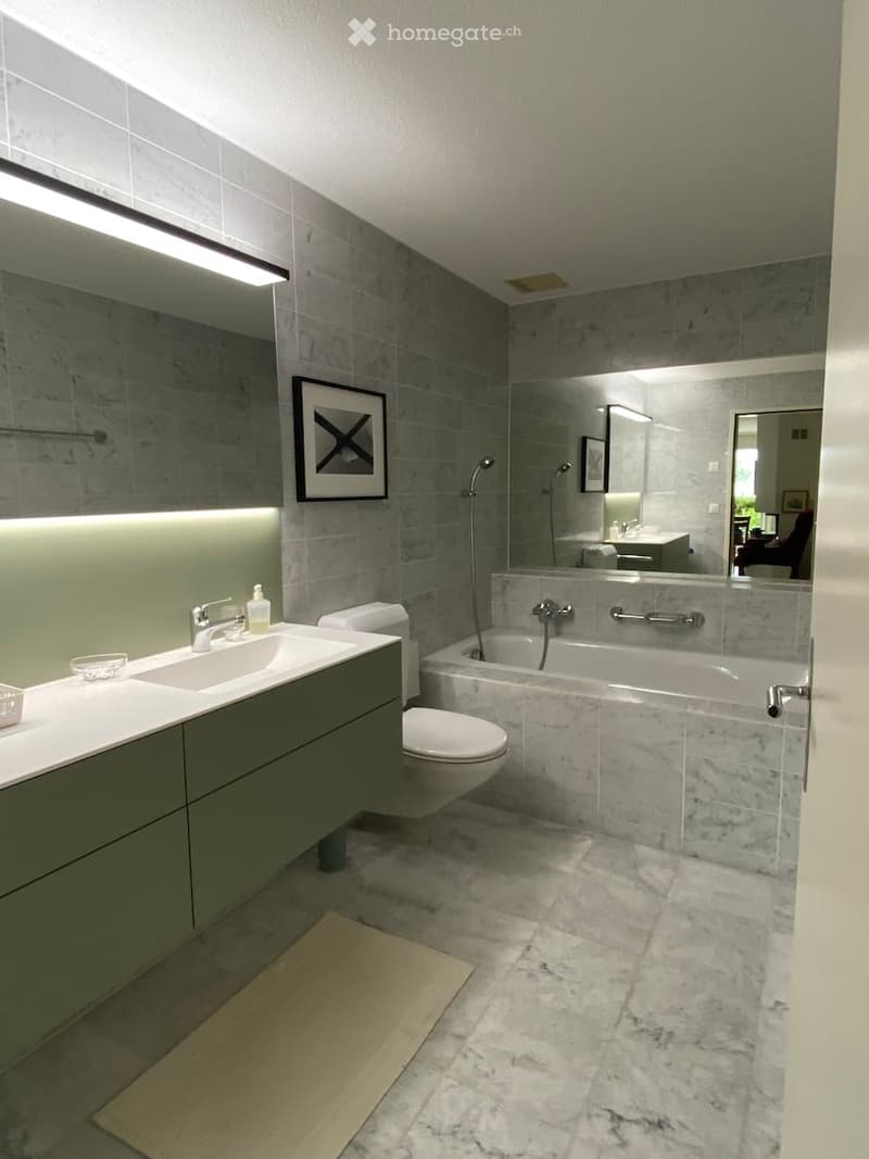 Modern bathroom with marble tiles, a large mirror, a bathtub, a toilet, a sink with a faucet, and a framed picture on the wall.