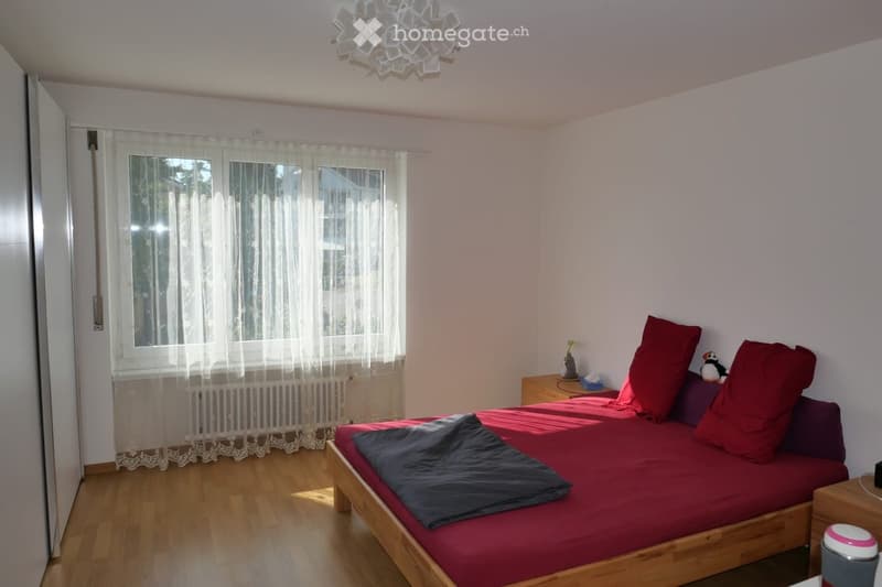A cozy bedroom with a wooden bed, red bedding, a nightstand, and a window with sheer curtains.